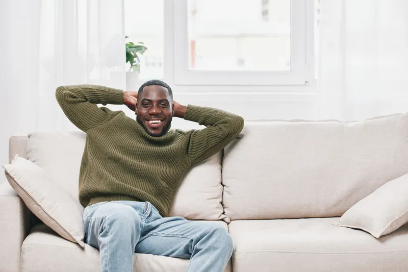 A joyful young Black man relaxes on a modern sofa, showcasing a casual style in a cozy indoor setting, wearing a green sweater and light jeans