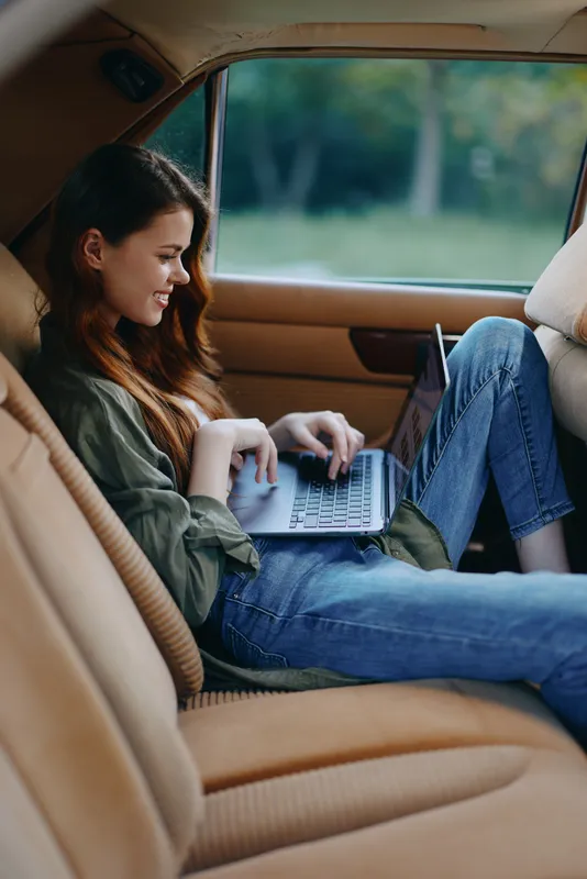 Relaxed young woman using laptop in vintage car interior, smiling and enjoying a peaceful moment in nature, with a focus on comfort and leisure.