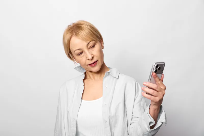 Elderly woman looks worried while holding a smartphone against a plain white background, with a tense posture and downward gaze. Concept for stress, loneliness, and mental health in modern digital
