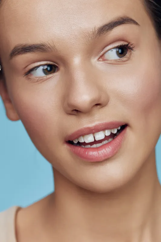 Close up portrait of a young woman with a joyful expression, showcasing natural beauty against a soft blue background, emphasizing her clear skin and radiant smile