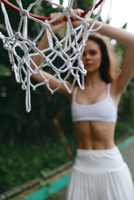 Athletic young woman in a white sports outfit with a basketball hoop, showcasing strength and confidence in a vibrant outdoor setting