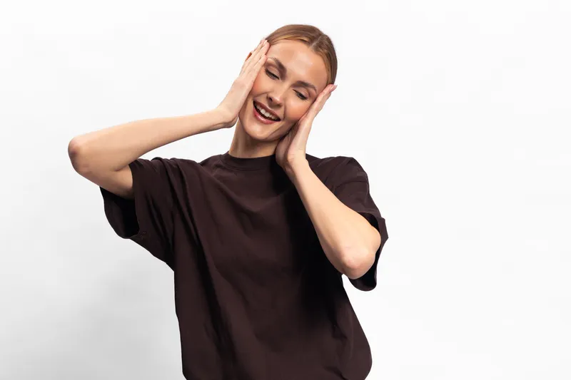 Smiling young woman in casual dark shirt touches face with hands, eyes closed, expressing joy and relaxation. Isolated on white background. Concept of happiness, calm, and positive emotions.