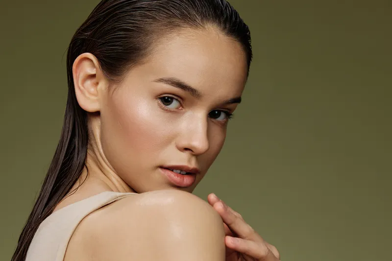 Young woman with wet hair, wearing a nude top, showcasing natural beauty and confidence against a soft green background