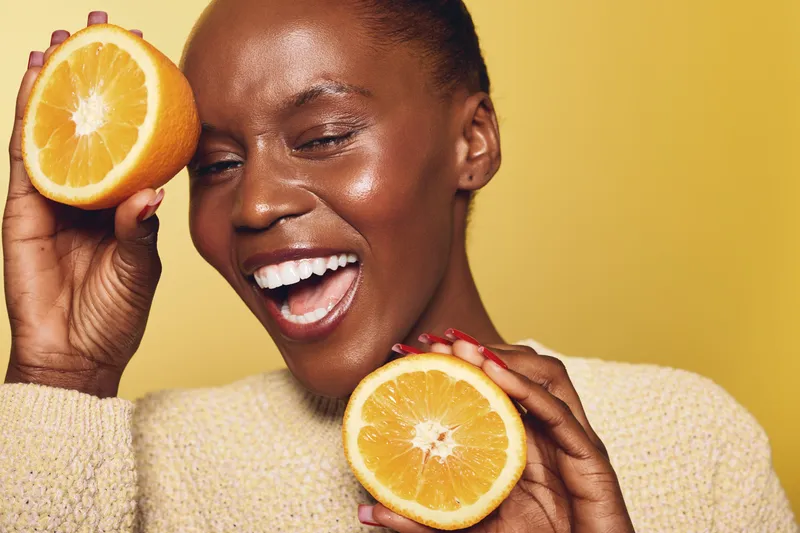 Happy African woman with dark skin holds two halves of fresh orange fruit near her face, smiling with closed eyes on yellow background. Concept of healthy lifestyle and natural vitamins.