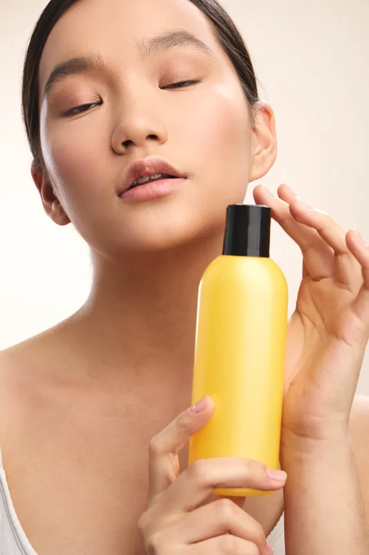 asian woman holds a yellow cosmetic bottle near her face with closed eyes, showcasing a calm skincare moment, soft studio lighting, and luminous smooth skin.