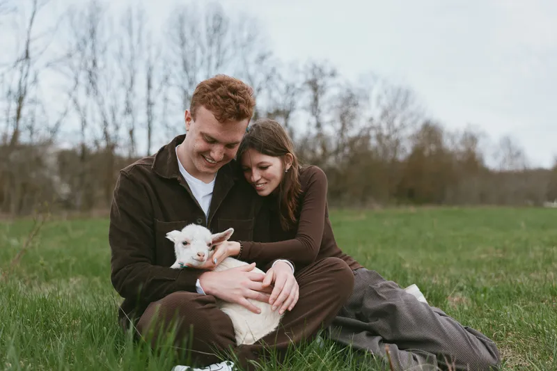 A couple sits on the grass, smiling as they cradle a white goat kid, enjoying a peaceful rural moment. A lifestyle scene focused on connection, care, and gentle animal companionship.