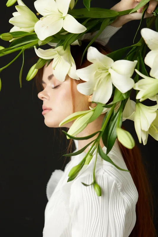 Young woman with long red hair adorned with white lilies, expressing serenity and elegance against a dark background Ideal for concepts of beauty and nature