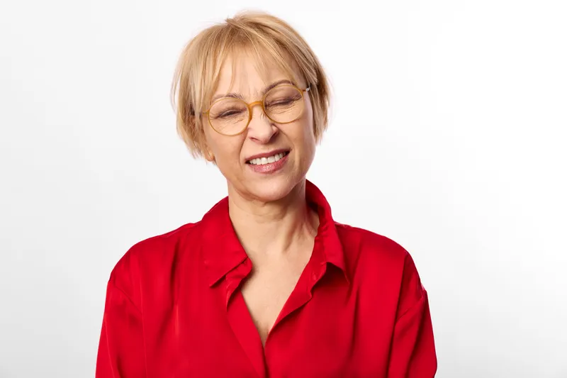 Senior woman portrait, blonde hair, glasses, warm smile in red blouse, studio white background, relaxed posture, soft light, closeup, calm expression