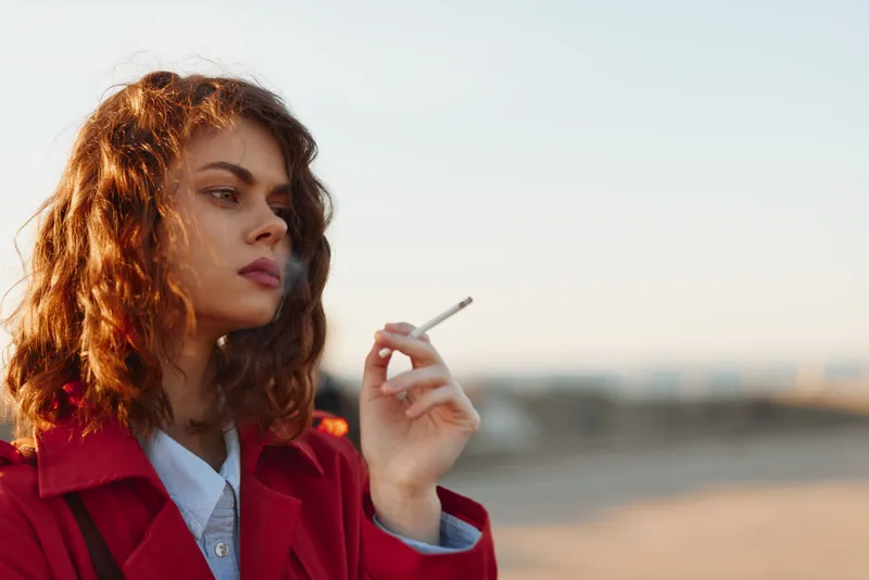 Young woman with curly red hair in a red jacket smokes a cigarette on a sunny beach with a distant boardwalk, the golden hour light highlighting a contemplative mood.