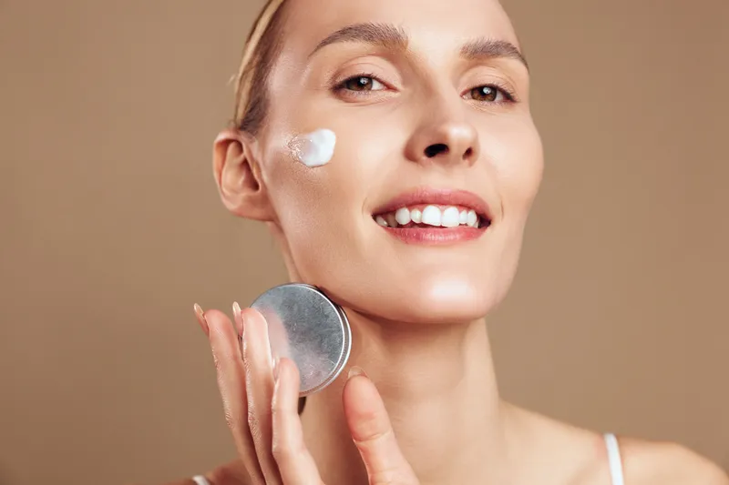 Smiling woman with clear skin applying white face cream on cheek, holding cosmetic jar. Close-up on beige background. Concept of skincare, beauty routine, and natural wellness.