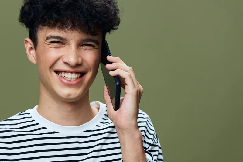 smiling man talking on smartphone in casual striped shirt with curly hair on olive background, expressing happiness and friendly communication, modern lifestyle