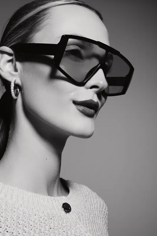Portrait of a fair skinned white woman wearing oversized geometric sunglasses and hoop earrings. She poses confidently against a plain grey background in monochrome style.