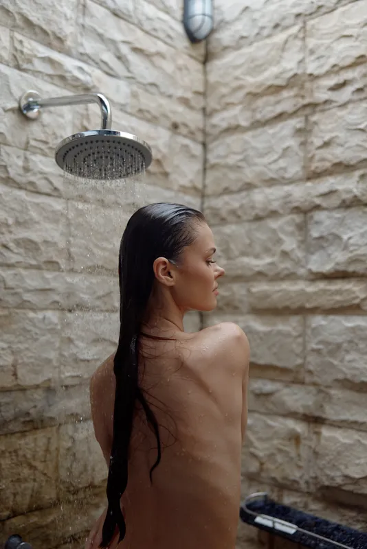 young woman showering outdoors with wet hair under rain showerhead on textured stone wall background in natural light