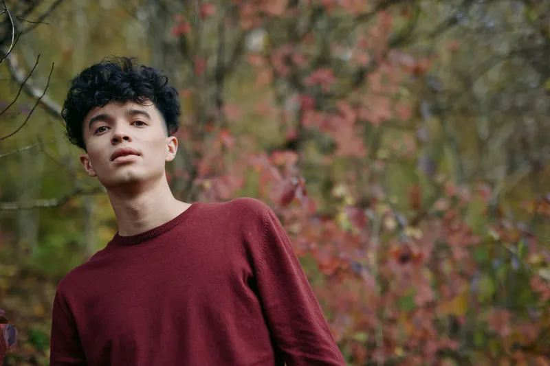 An authentic portrait of a young man in a burgundy sweater, standing in an autumnal grove with soft light, conveying real emotion and genuine self expression.