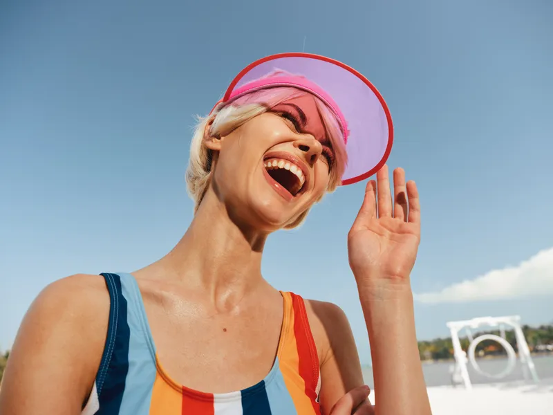 Joyful woman on a sunny beach wears a colorful striped swimsuit and pink visor, laughing with head tilted back as bright sky frames her carefree summer moment.