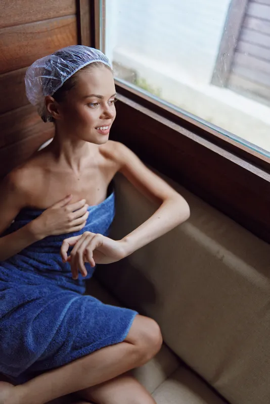 Relaxed woman in blue towel wearing shower cap sits by window in wooden spa room, enjoying natural light and peaceful moment of self-care and wellness.