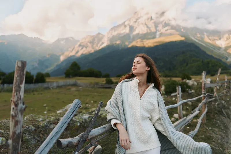 Bright outdoor moment with a woman embracing nature, casual lifestyle portrait, mountain scenery, soft breeze, relaxed pose, light colors, rustic wooden fencing backdrop