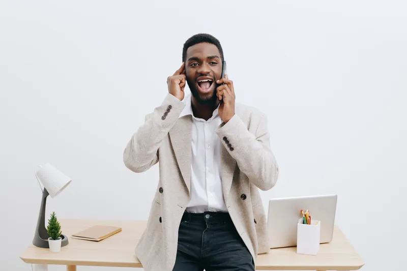 Smiling man talking on mobile phone in office, wearing smart casual outfit, standing at desk with laptop, plant and stationery, light background. People lifestyle concept.