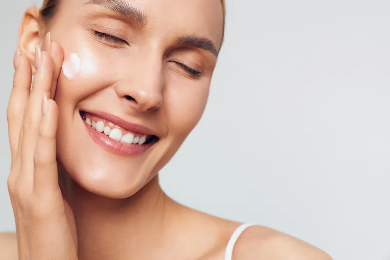 A young woman applies cream to her cheek in a bright studio, smiling softly as a skincare routine moment unfolds, conveying calm self care and gentle daytime pampering