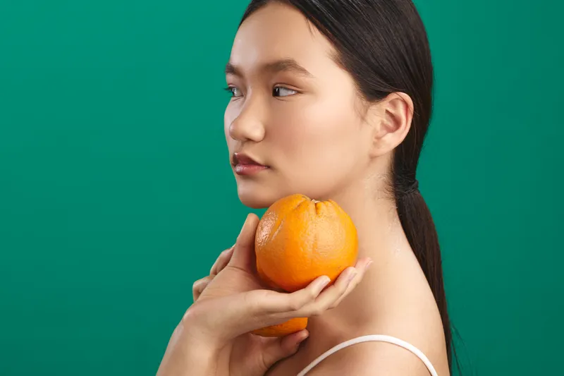 asian woman holds orange near her face against a solid green background, calm expression and soft lighting, creating a fresh, minimalist portrait with a natural, organic feel.