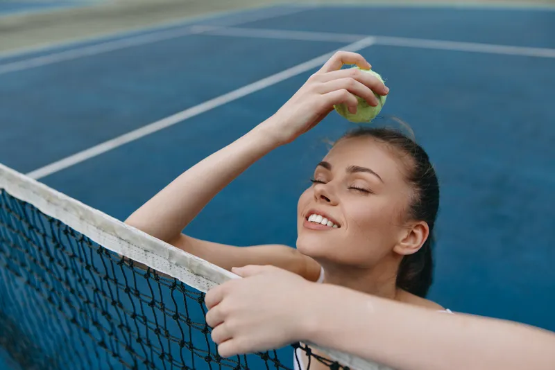 Young female tennis player smiling while holding a tennis ball against a blue court, showcasing joy and athleticism in sportswear