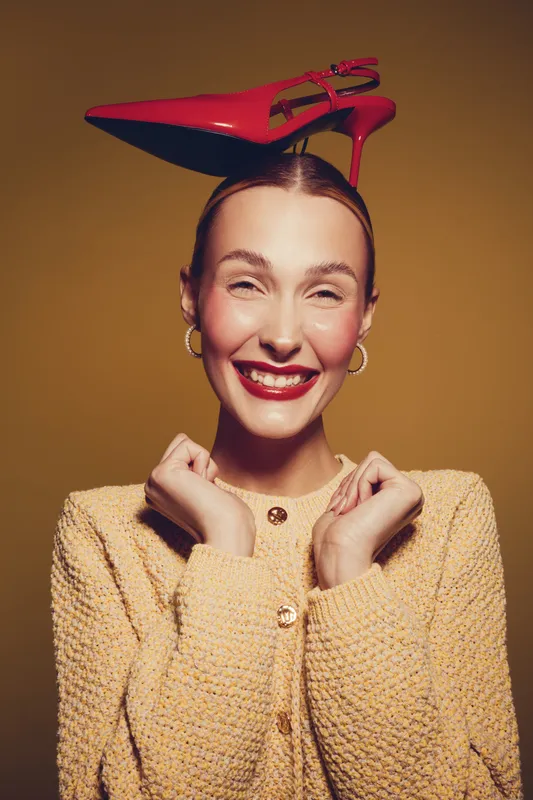 Smiling woman in yellow sweater balances red high heel shoe on her head, showing playful mood and fashion concept with bright studio lighting on brown background.