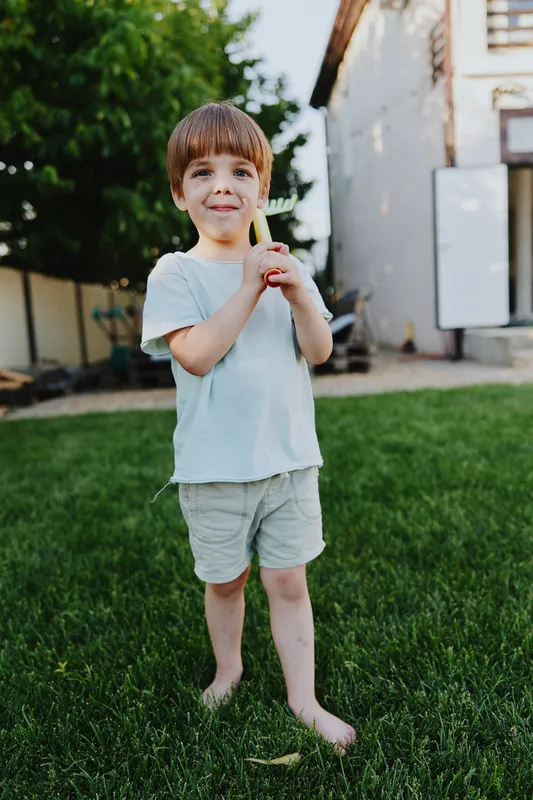 Happy boy standing barefoot on green grass, holding a colorful toy. Natural light highlights the joyful moment in a relaxed outdoor setting.
