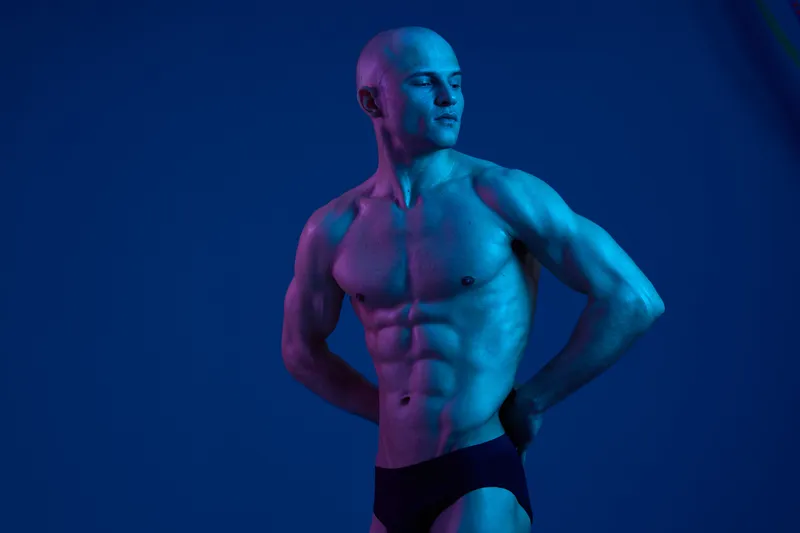Strong man posing confidently, showcasing muscular physique against a vibrant blue background with creative lighting, emphasizing the power of the human body.