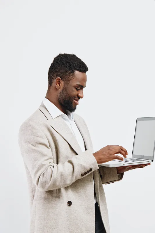 Smiling man in beige blazer using laptop on plain white background, casual business style, professional appearance, focused expression, modern clothing, studio portrait.