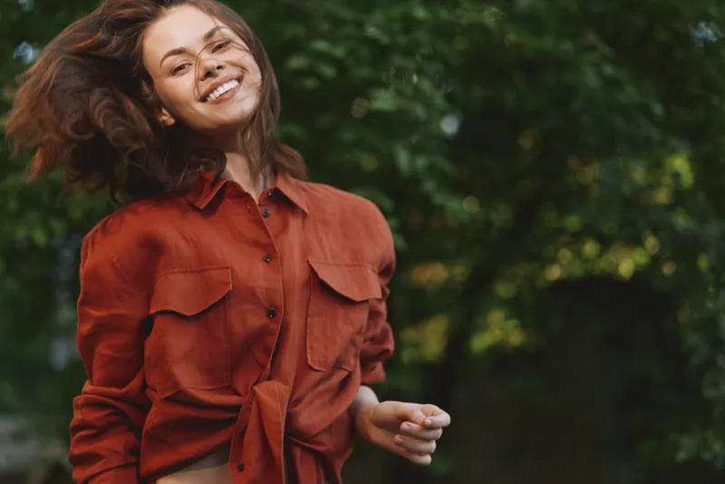 Smiling woman in burnt orange shirt outdoors, candid lifestyle portrait, natural light, park setting, relaxed mood, casual fashion, happiness, sunlight energy