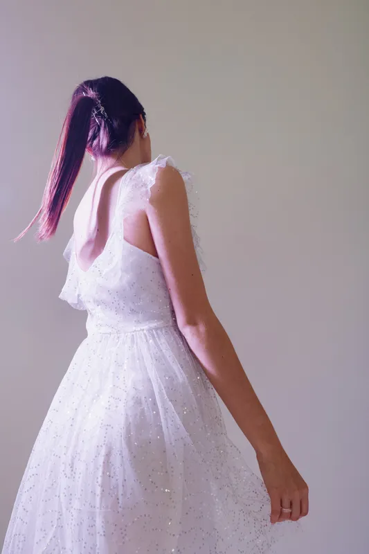 Young woman in a white elegant dress with a low back poses with her back to the camera. Soft lighting highlights the texture and delicate details of the fabric. Concept of beauty fashion.