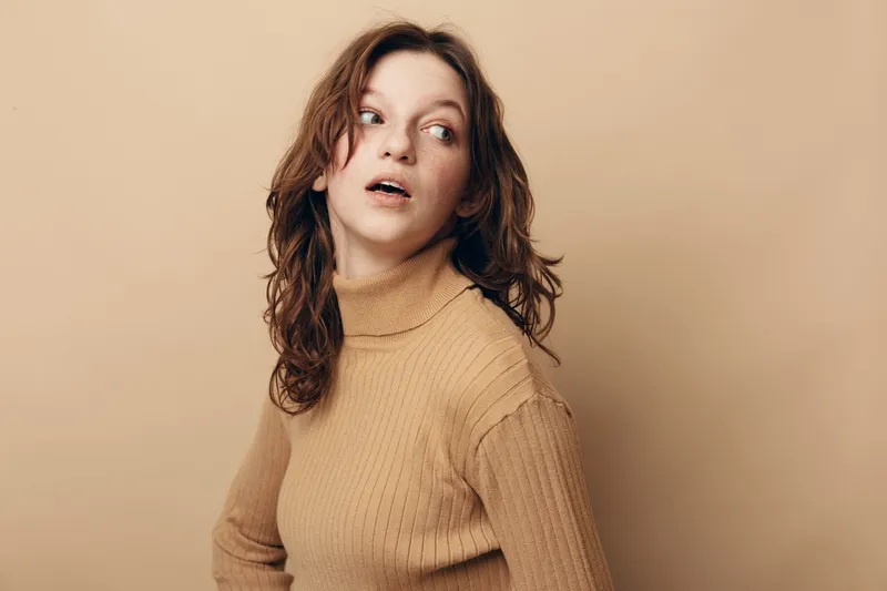 Young woman with curly hair wearing beige turtleneck looks sideways with surprised expression. Studio portrait on neutral background. Concept of curiosity, emotion, and casual fashion.