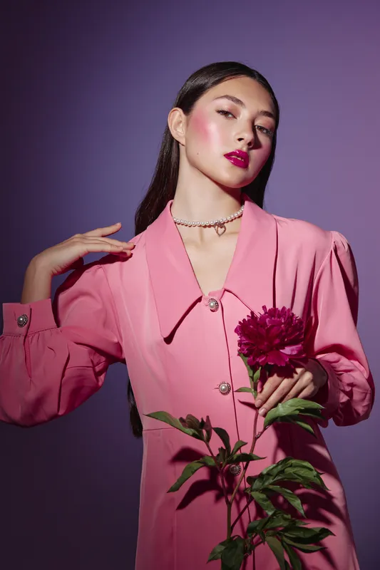 Fashion model with long hair posing elegantly in a pink dress holding dark red flower. Perfect lighting enhances vibrant makeup and stylish self-assured expression. Beauty concept.