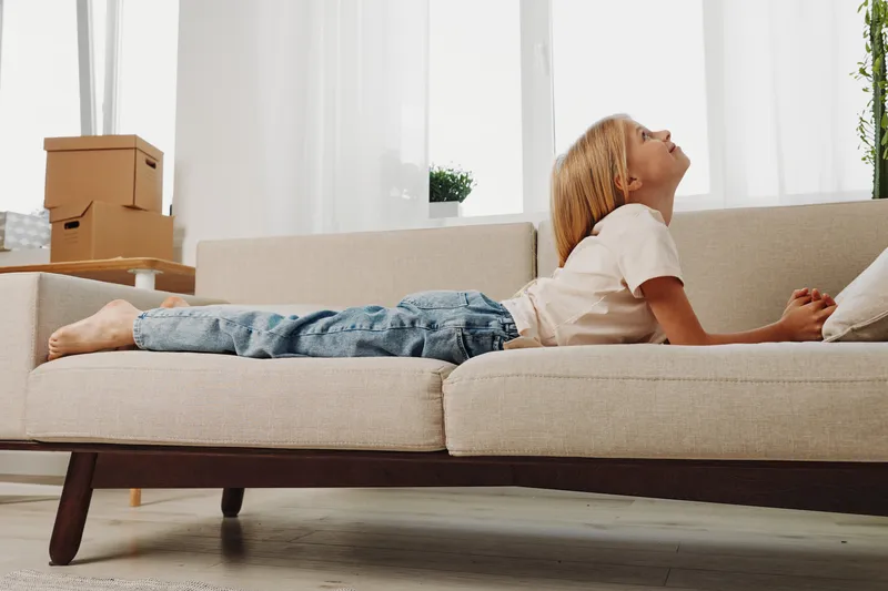 Young boy with blonde hair lying on beige sofa in bright living room, looking up with calm expression. Cozy home interior with natural light and casual outfit.