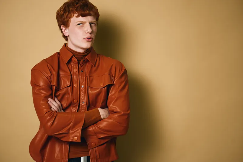 Young man with red hair wearing a brown leather jacket and turtleneck sweater standing with crossed arms against beige background with thoughtful expression.