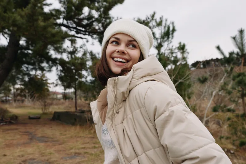 Woman outdoor winter jacket smiling park portrait, cold weather fashion in a casual moment outdoors with a beanie, capturing a natural lifestyle scene in soft daylight and relaxed mood.