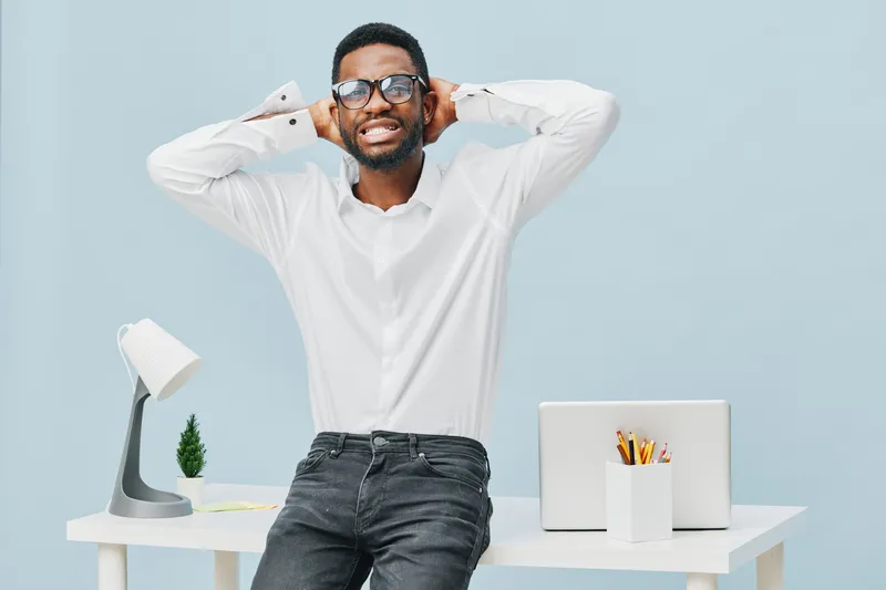 Joyful young man with glasses standing in modern office workspace, smiling and stretching hands behind head, casual white shirt and black jeans, relaxed mood, minimalistic decor, bright background, li