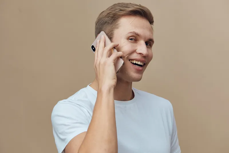 Smiling man talking on mobile phone with cheerful expression, casual light blue t-shirt, beige background, close-up studio portrait. People lifestyle concept