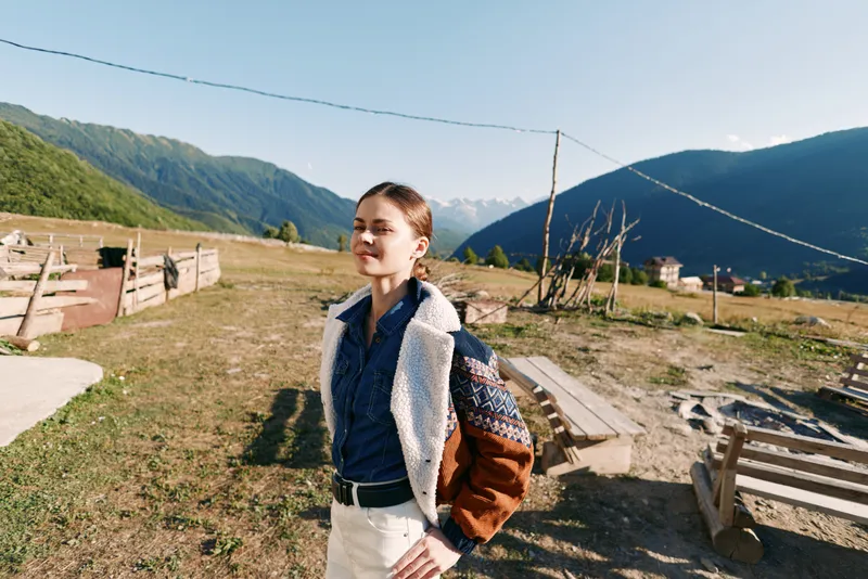 Woman in mountains village standing outdoors with backpack and jacket, sunlight on face and eyes closed enjoying nature and travel, casual style in peaceful rural landscape.