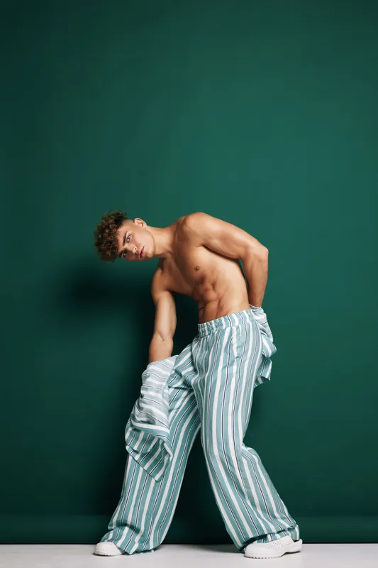 Young man posing shirtless with striped pants and jacket against dark green wall. Fashion, style and model with athletic build in casual outfit on white floor in studio.