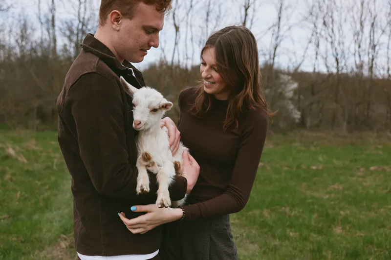 A couple outdoors hold a small goat kid in a grassy field, sharing a warm moment. Natural light highlights care, family bonds, and simple rural living in a candid lifestyle scene that feels intimate.