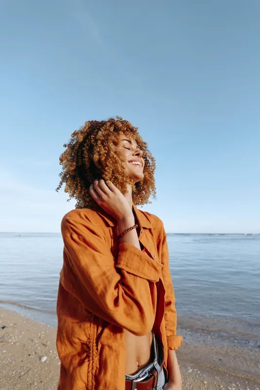 Young woman with curly hair smiles on a sunny beach in an orange shirt, feeling confident.