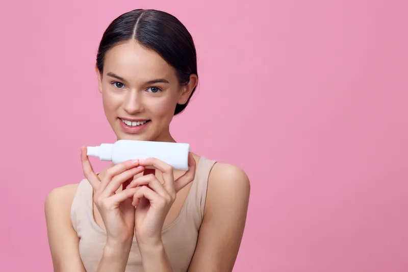 Young woman with a skincare cream smiling against a pink background, showcasing a healthy skincare routine and self care, perfect for beauty and wellness themes