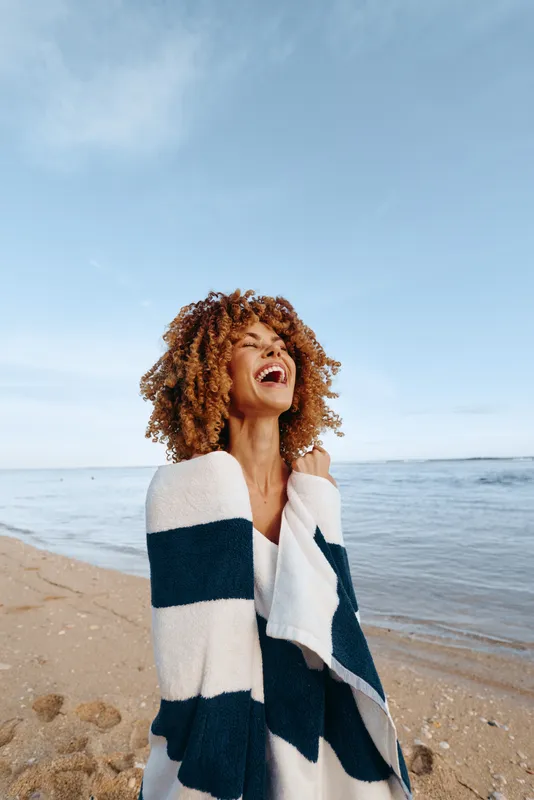 Young woman with curly hair smiles on a sunny beach, wrapped in a striped blanket as gentle waves roll in behind her, creating a carefree, relaxed mood.
