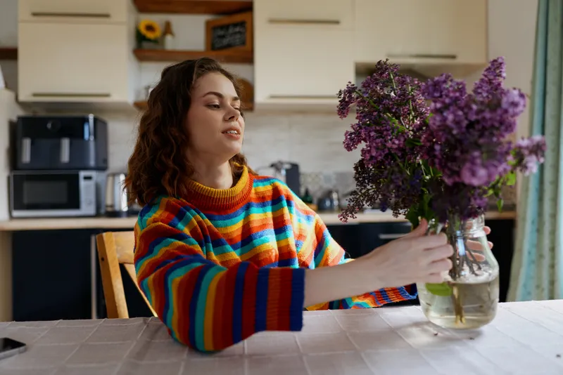 A young woman with curly hair sits at a kitchen table in a colorful striped knit sweater, admiring a bouquet of purple flowers in a glass vase.
