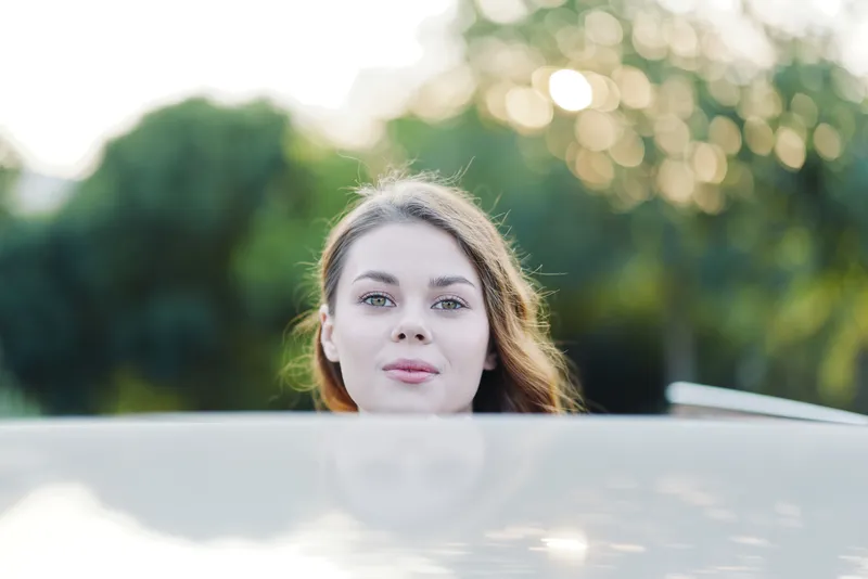 Young woman with natural beauty looking over a car hood, capturing a soft and serene moment in a lush green background, ideal for lifestyle and travel themes.