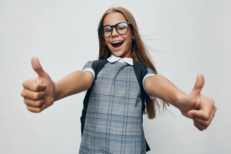Girl with glasses showing thumbs up in school uniform with backpack smiling standing against white background. Cheerful child expressing positivity and confidence indoors.