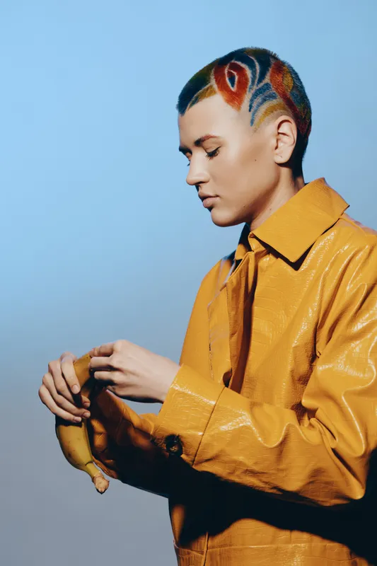 Woman with colorful short hair and artistic shaved patterns wearing yellow textured coat holds a banana against a soft blue background in creative studio portrait.