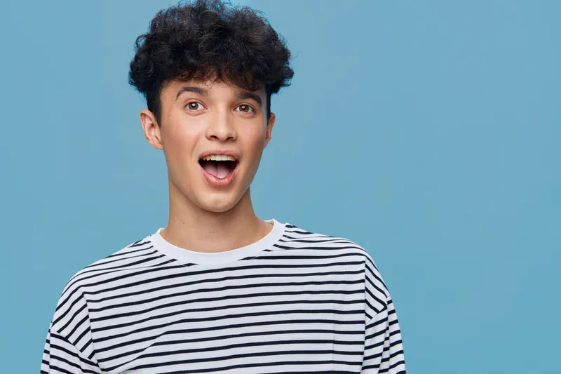 surprised young man with curly hair wearing a striped t-shirt against blue background expressing shock and excitement in casual style