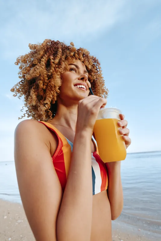 Happy young woman with curly hair drinks orange juice from a glass with straw on the beach under blue sky, wearing colorful swimsuit, enjoying summer vacation and healthy lifestyle